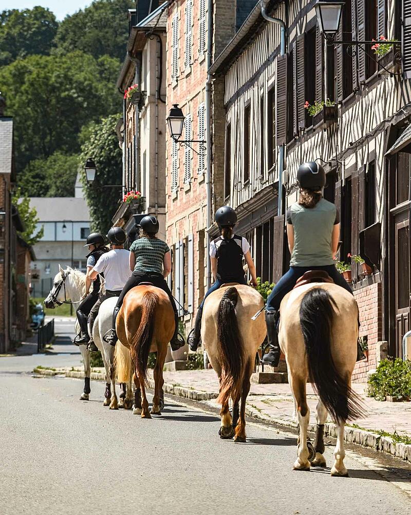 A group of riders on horseback walking down a village street lined with classic half-timbered stone houses, an outdoor activity proposed near your Les Castels camping.