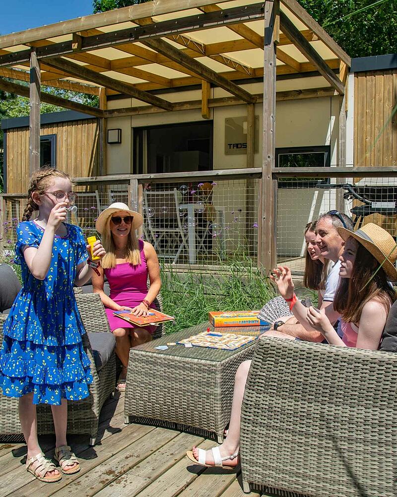 A family enjoys quality time together playing a board game and blowing bubbles on the spacious wooden terrace of a premium, modern mobile home at a Les Castels campsite.