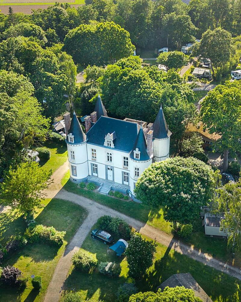 Drone aerial view of a small white chateau with pointed roofs surrounded by a lush green park and campsite pitches in Saint-Malo.