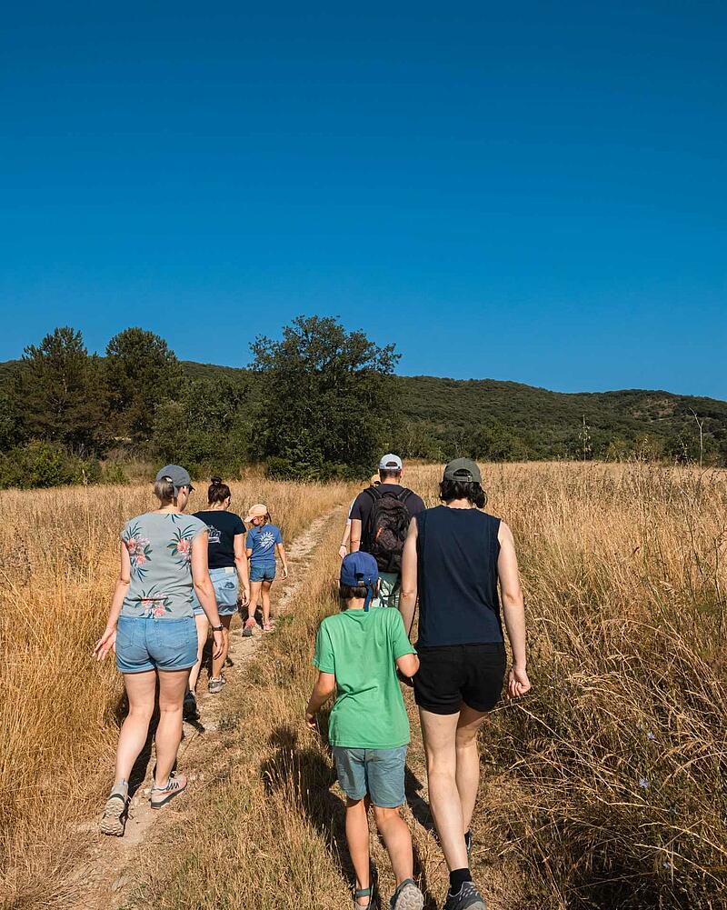A multi-generational family of six hikes together on a narrow dirt path through a grassy field under a clear blue sky at a Les Castels camping estate.