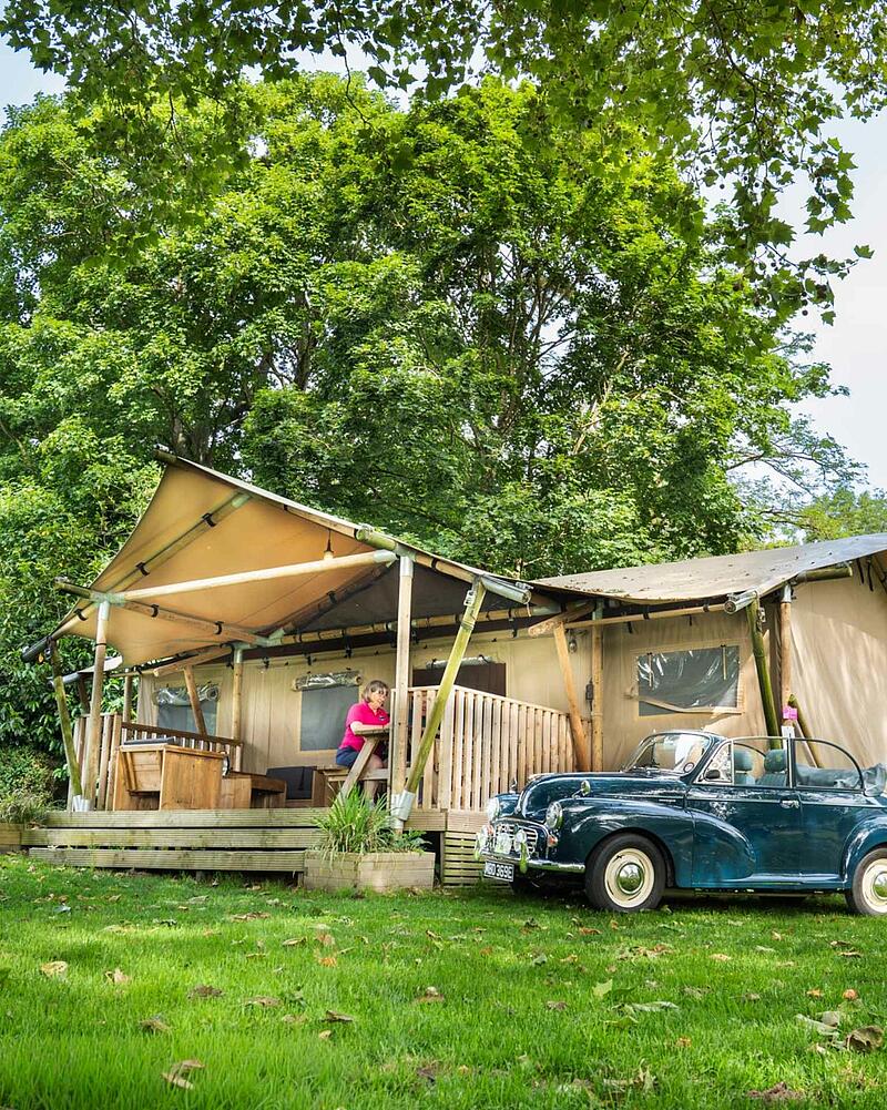 A large canvas glamping safari tent with a wooden porch set amongst lush green trees on a premium pitch at a Les Castels 5-star campsite.