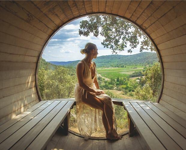 Panoramic barrel sauna experience at Castels Domaine de Sévenier, Ardèche, France. A woman relaxes inside a wooden barrel sauna looking out at the green valley at Castels camping Domaine de Sévenier.