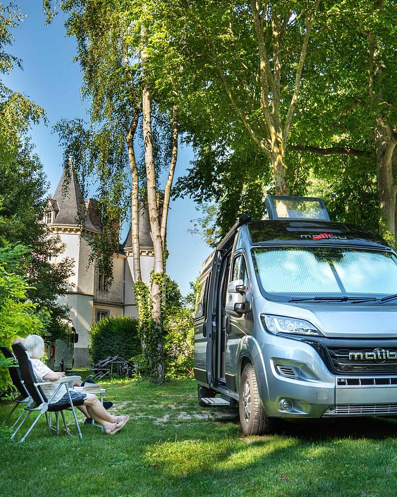 A senior couple sitting on camping chairs in the shade of trees next to their Malibu campervan, with the Ville Huchet chateau visible in the background.