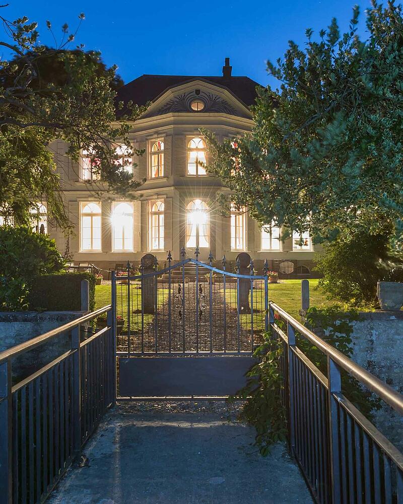 The main manor house of Camping Les Castels La Bien-Assise beautifully illuminated at night, viewed from across a small bridge and iron gate under a clear evening sky.