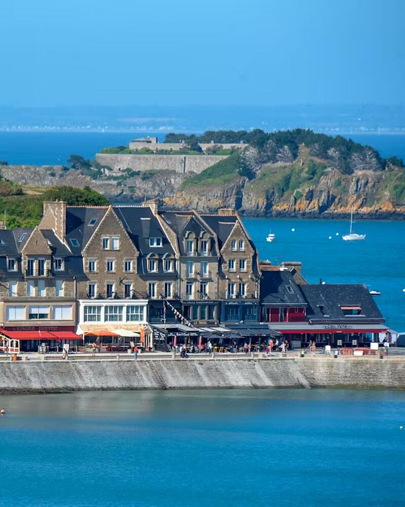 View of the stone facades of hotels and restaurants on the port of Cancale in Brittany, with the fort of Rimains Island in the distance on a turquoise sea.