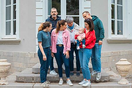A multi-generational family of adults, seniors, and a baby standing on the stone steps outside a classic French estate building, talking and smiling together.