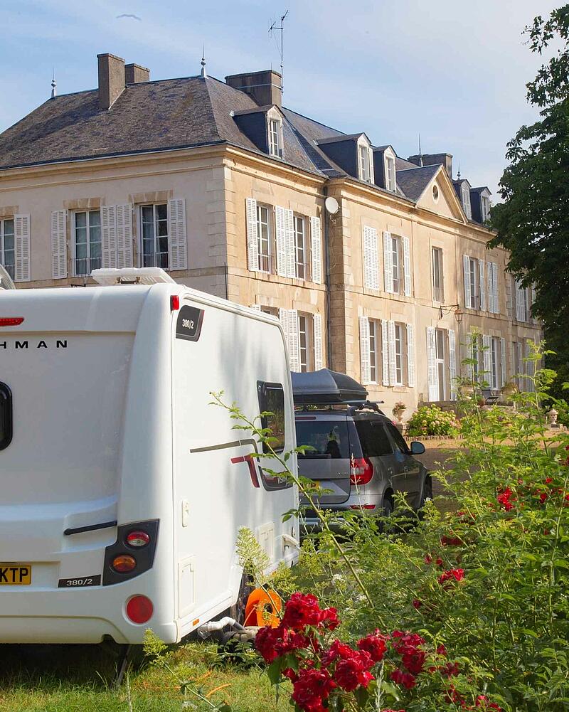 The back of a white caravan and a grey SUV parked on a sunny grass pitch in front of a grand, historic stone chateau, beautifully framed by blooming red roses in the foreground.