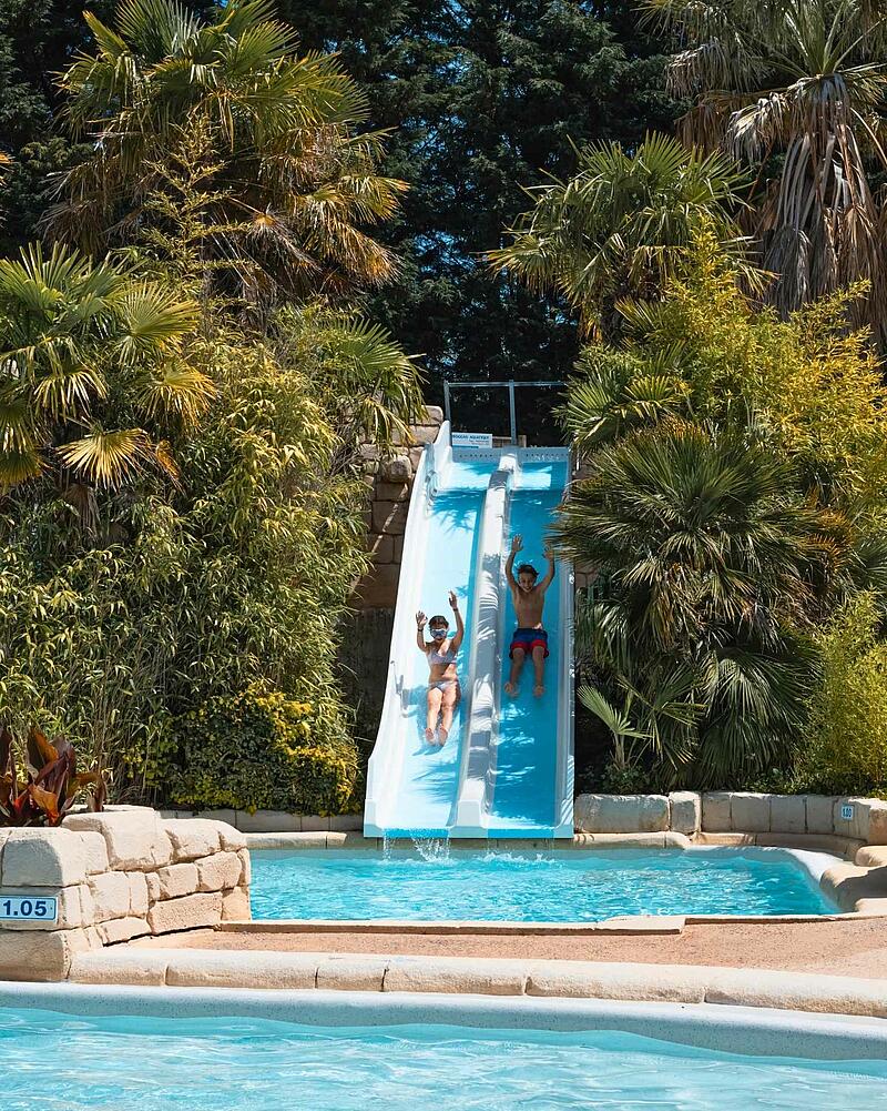 Two kids cheering with raised hands while sliding down a blue double-lane water slide surrounded by palm trees at a Les Castels family campsite.