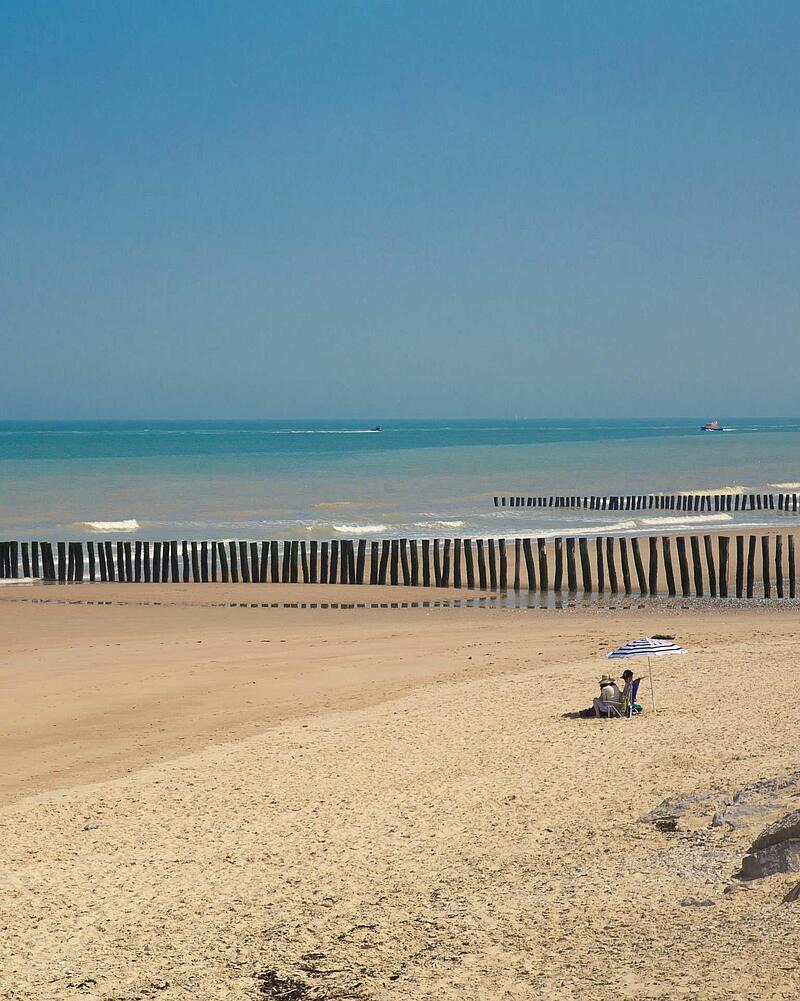 Wide sandy beach in France with rows of vertical wooden groynes leading into the blue ocean under a clear sky, typical of a seaside camping landscape.