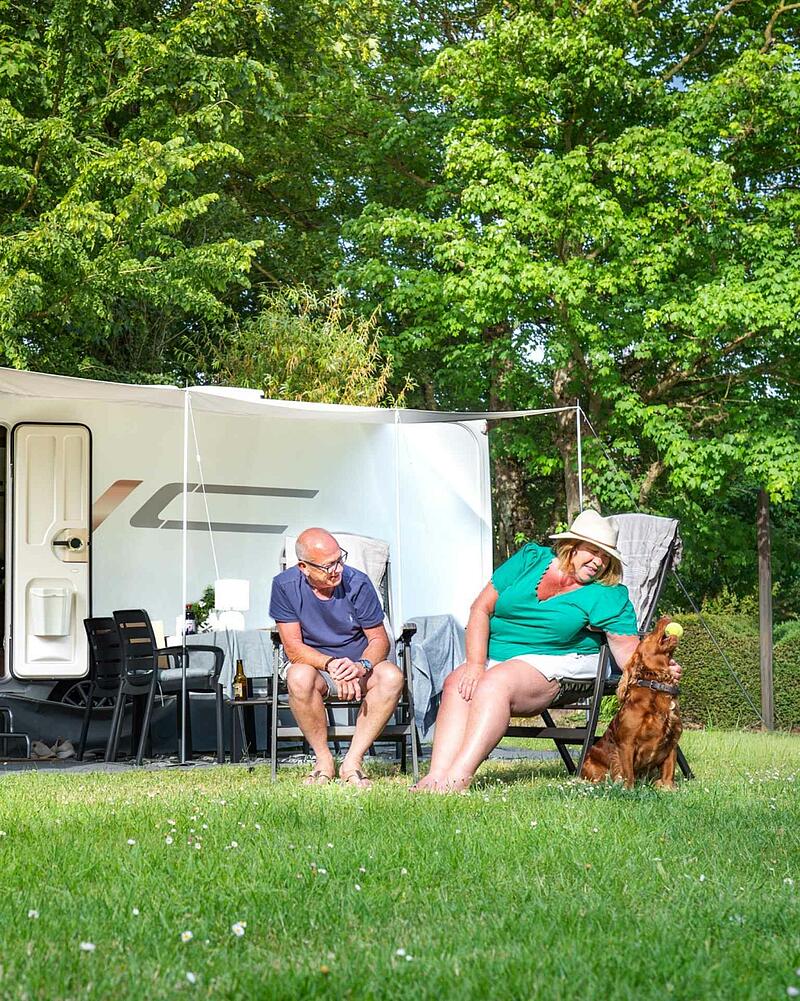 A man and woman relaxing in chairs outside their caravan on a lush green lawn, playing with their brown dog holding a tennis ball at a pet-friendly campsite.