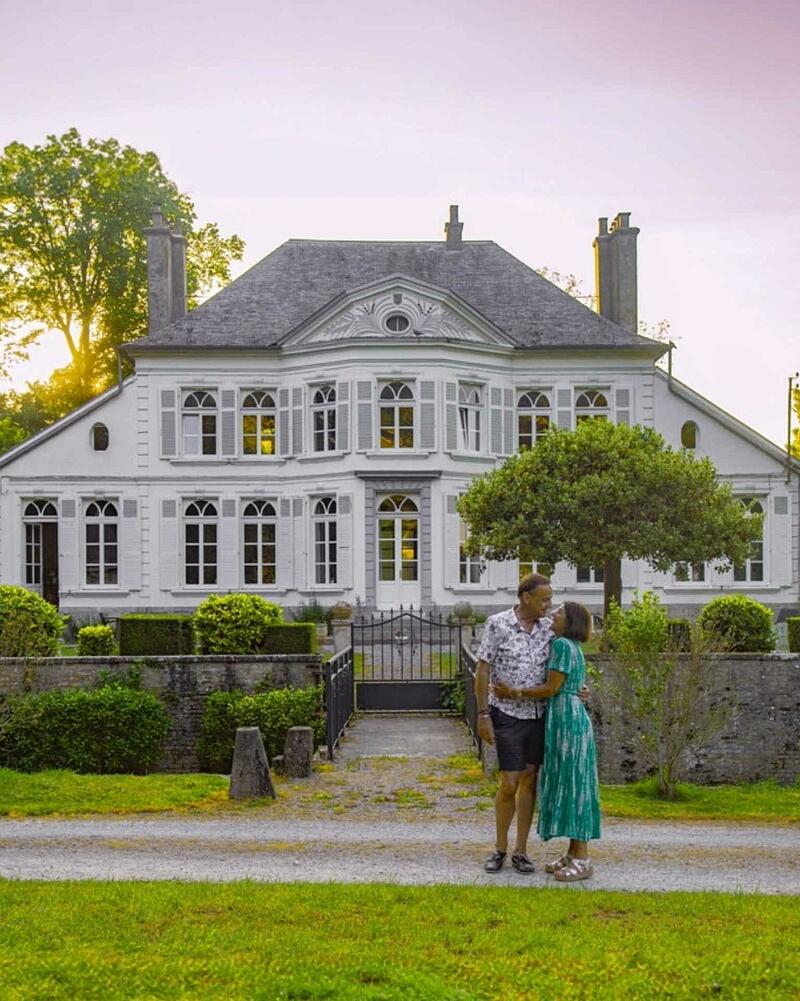 A couple standing lovingly in front of a historic French chateau estate at Camping La Bien-Assise during a warm, golden sunset.