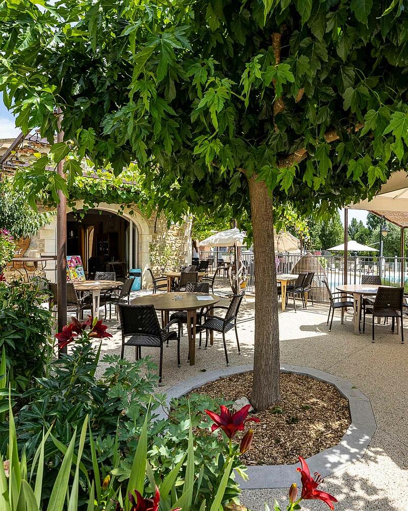 Outdoor dining terrace with tables and chairs under lush green trees at Les Castels Domaine de Sévenier campsite near Ardèche chalet rentals.
