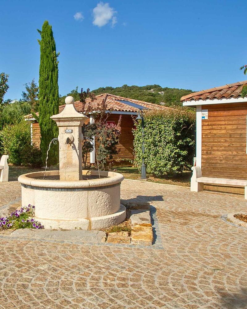 Stone fountain in a stone-paved square surrounded by wooden chalets and Mediterranean greenery at Les Castels Domaine de Sévenier in Ardèche, France.