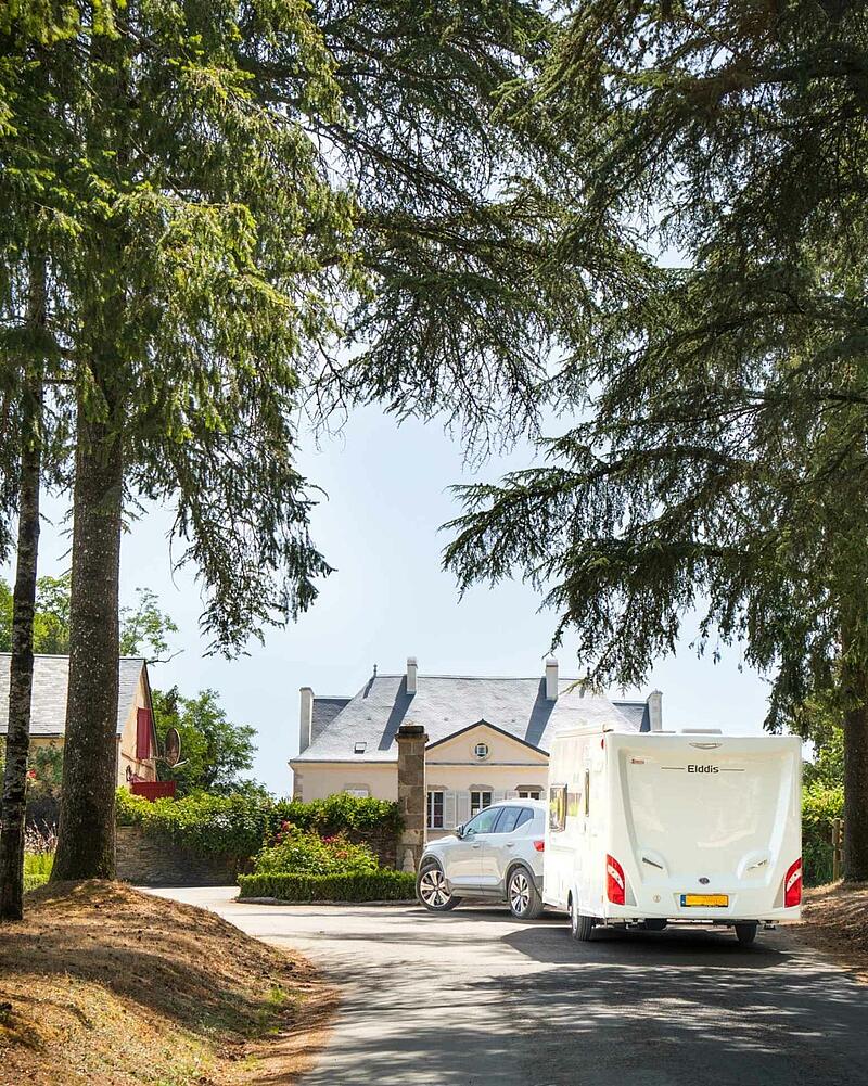 A stylish car towing a modern caravan is parked on a spacious pitch near a beautiful manor house, representing the high-end caravan spots ready for a spontaneous getaway at Les Castels