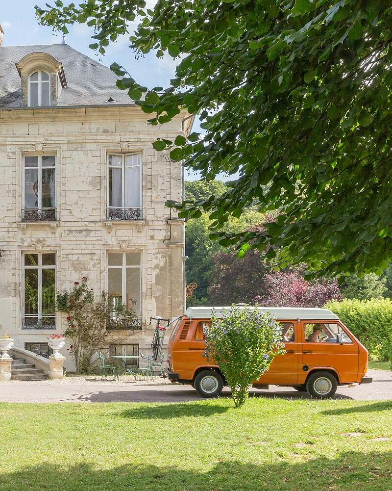 A classic orange campervan parked on the grass beside a majestic, historic stone chateau building, perfect for a road trip stopover from Dover to Normandy.