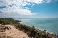 A scenic coastal view in Normandy featuring a dirt pathway on a grassy cliff overlooking a vast expanse of turquoise sea under a blue sky with fluffy white clouds, perfect for an outdoor stay at Les Castels.